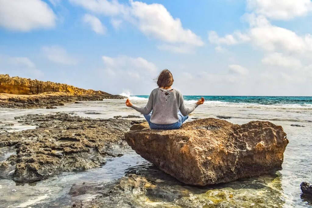 Frau sitzt im Lotussitz auf einem Felsen am Meer – Symbol für innere Ruhe, Achtsamkeit und Selbstregulation des Nervensystems.